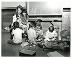 Children sitting on floor playing next to teacher by Indiana State University, University Archives